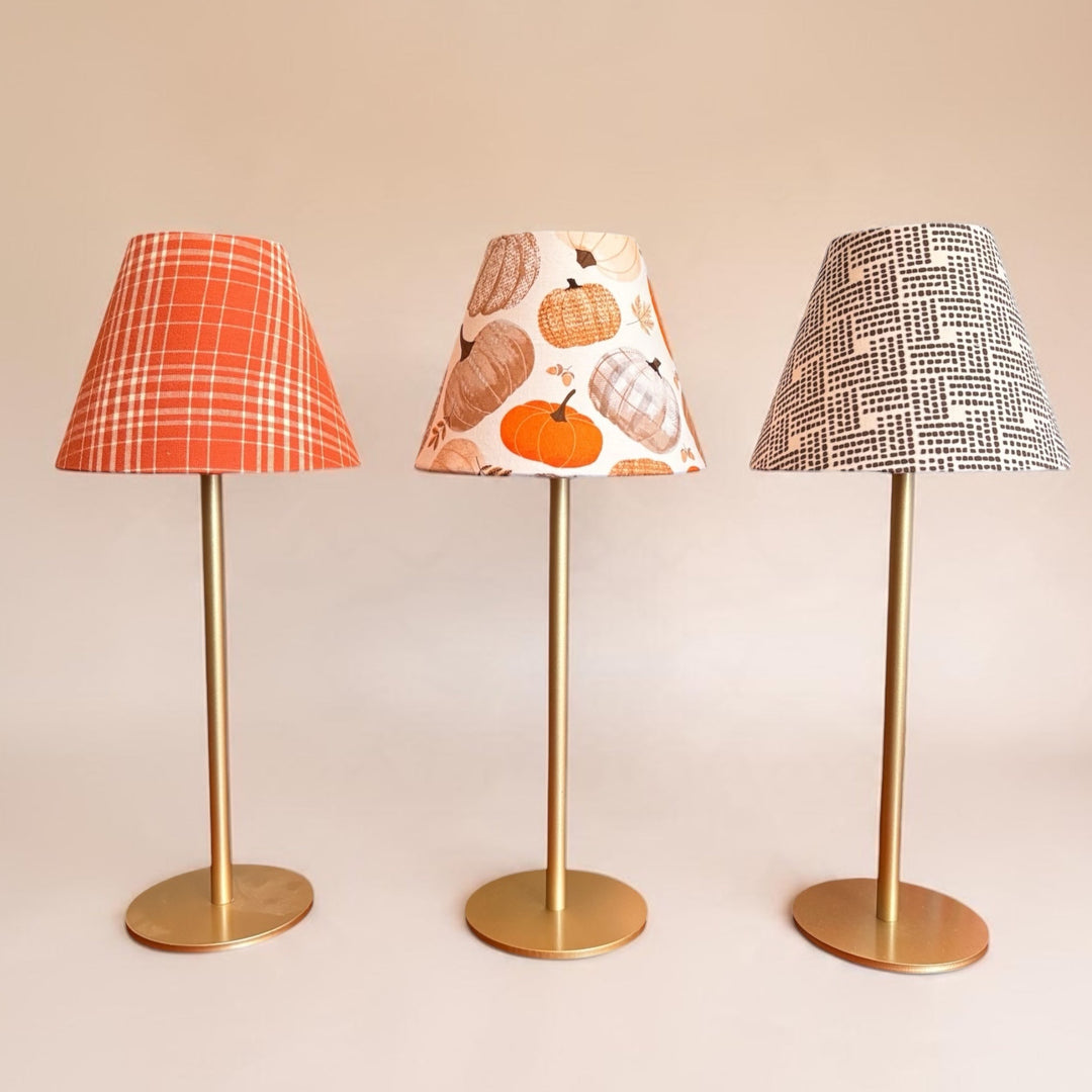 Three table lamps with slim gold metal stands and round bases, each with a patterned lampshade. The left lampshade is orange and white plaid, the middle lampshade has a pumpkin and shell pattern on a cream background, and the right lampshade has a black and white basketweave pattern.